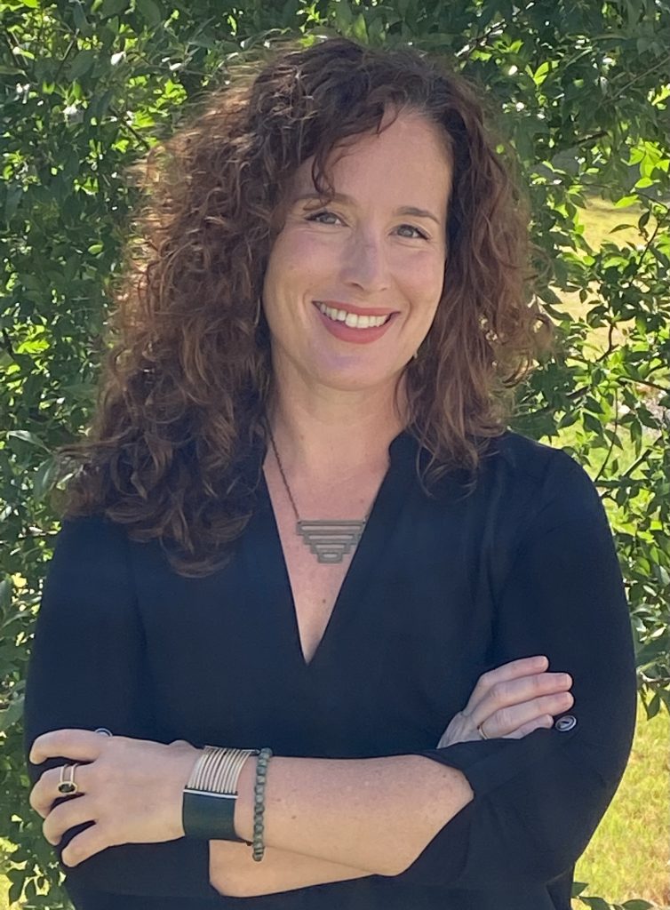 SMYAL's Executive Director Erin Whelan poses for a headshot. She is standing with her arms corssed in front of a very green tree. She has curly hair that hits just about shoulder length. She smiles while wearing a blue v-neck blouse and geometric necklace.