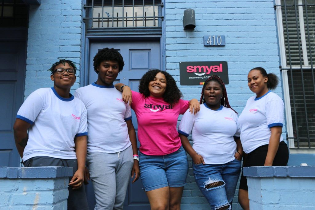 Group of BIPOC youth standing outside SMYAL's offices. There are five youth in total, the two on either side of the group are wearing white shirts, the youth in the middle is wearing a pink SMYAL shirt.