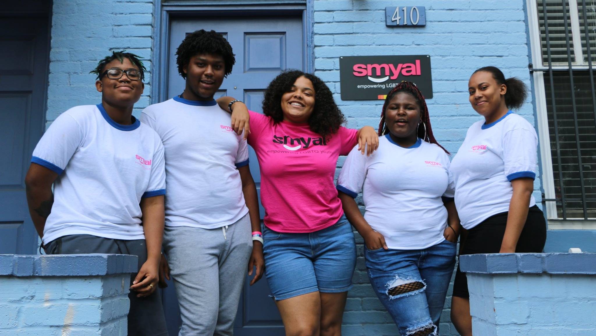 Group of BIPOC youth standing outside SMYAL's offices. There are five youth in total, the two on either side of the group are wearing white shirts, the youth in the middle is wearing a pink SMYAL shirt.